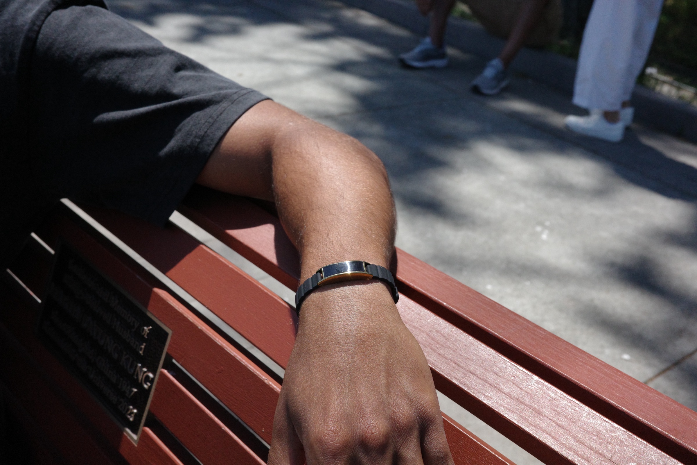 An arm rests over a red park bench in the sun, wearing Ray in Sunlit Gold on a black band